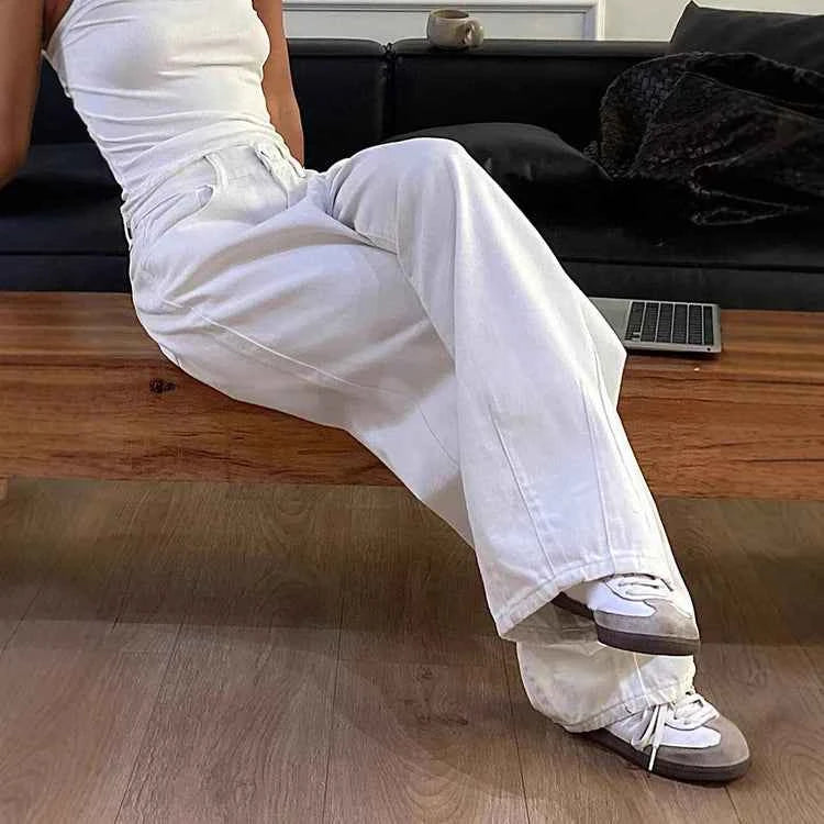 Woman wearing white wide-leg jeans and sneakers, seated on wooden bench in modern living room